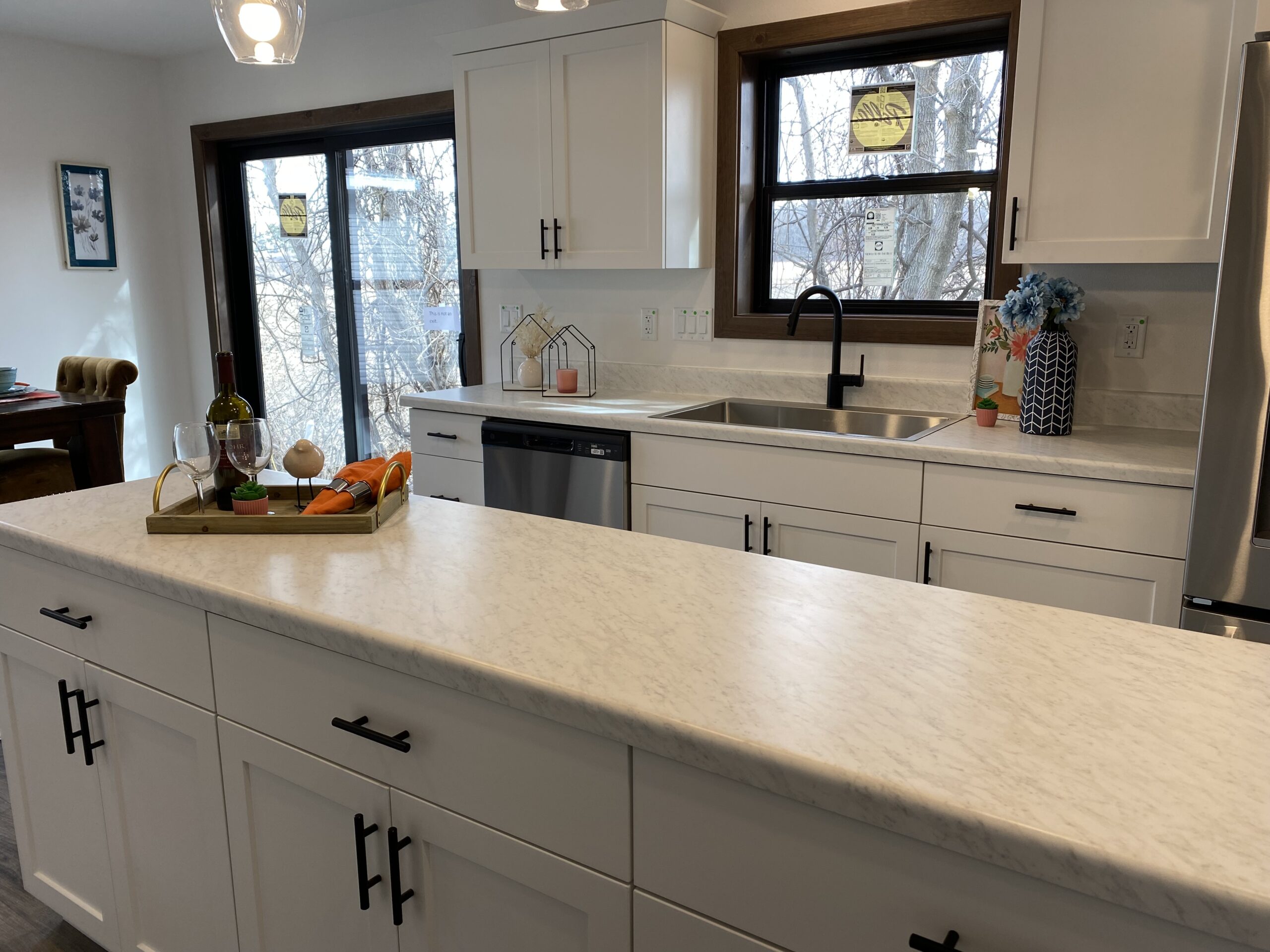 Modern kitchen with white cabinets and black handles, marble countertop, sink, and tray with wine, fruits. Bright ambiance with window view.