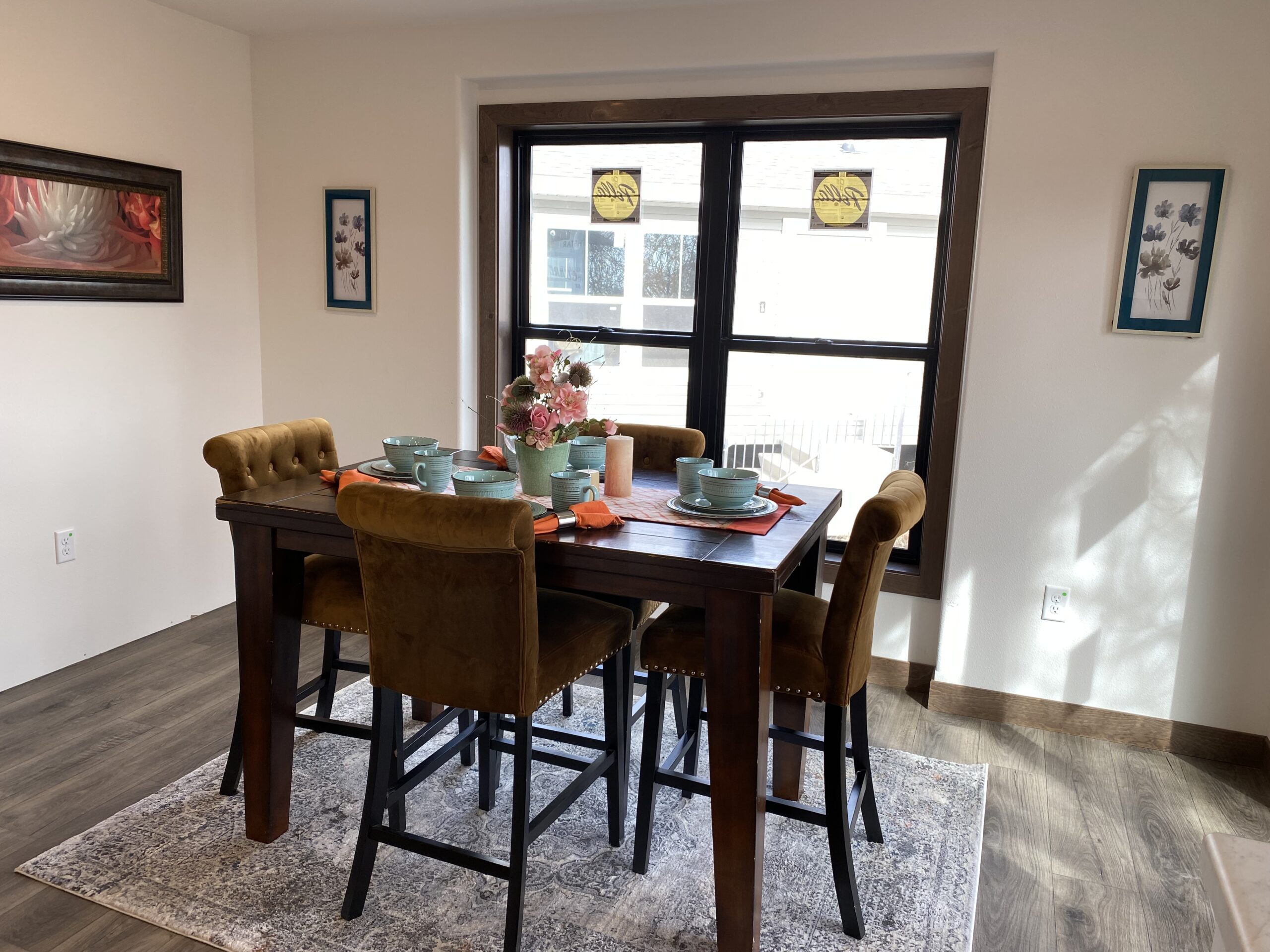 A cozy dining area with a dark wooden table set for four. Turquoise dishes and an orange runner complement the warm-toned chairs. Sunlight filters through a large window, enhancing the inviting atmosphere. Framed art and flowers add a touch of elegance.