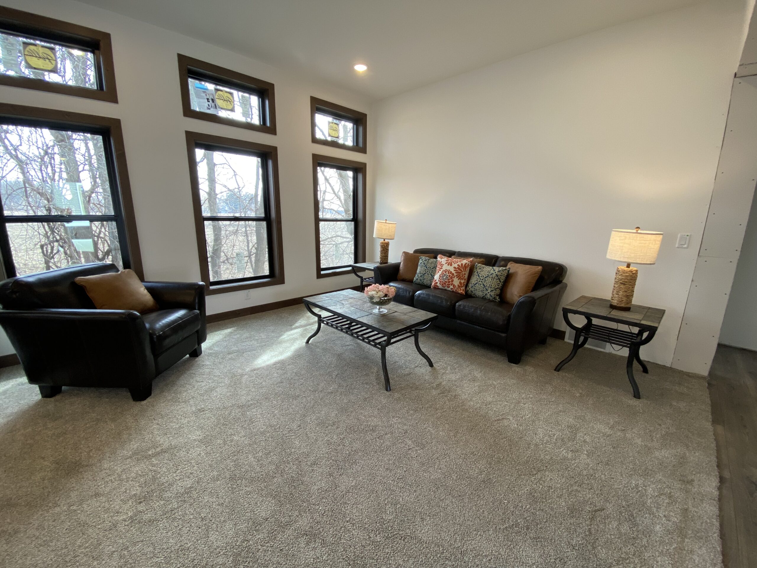 A cozy living room with large windows, a dark leather sofa, an armchair, a glass coffee table, and lit lamps creating a warm, inviting atmosphere.