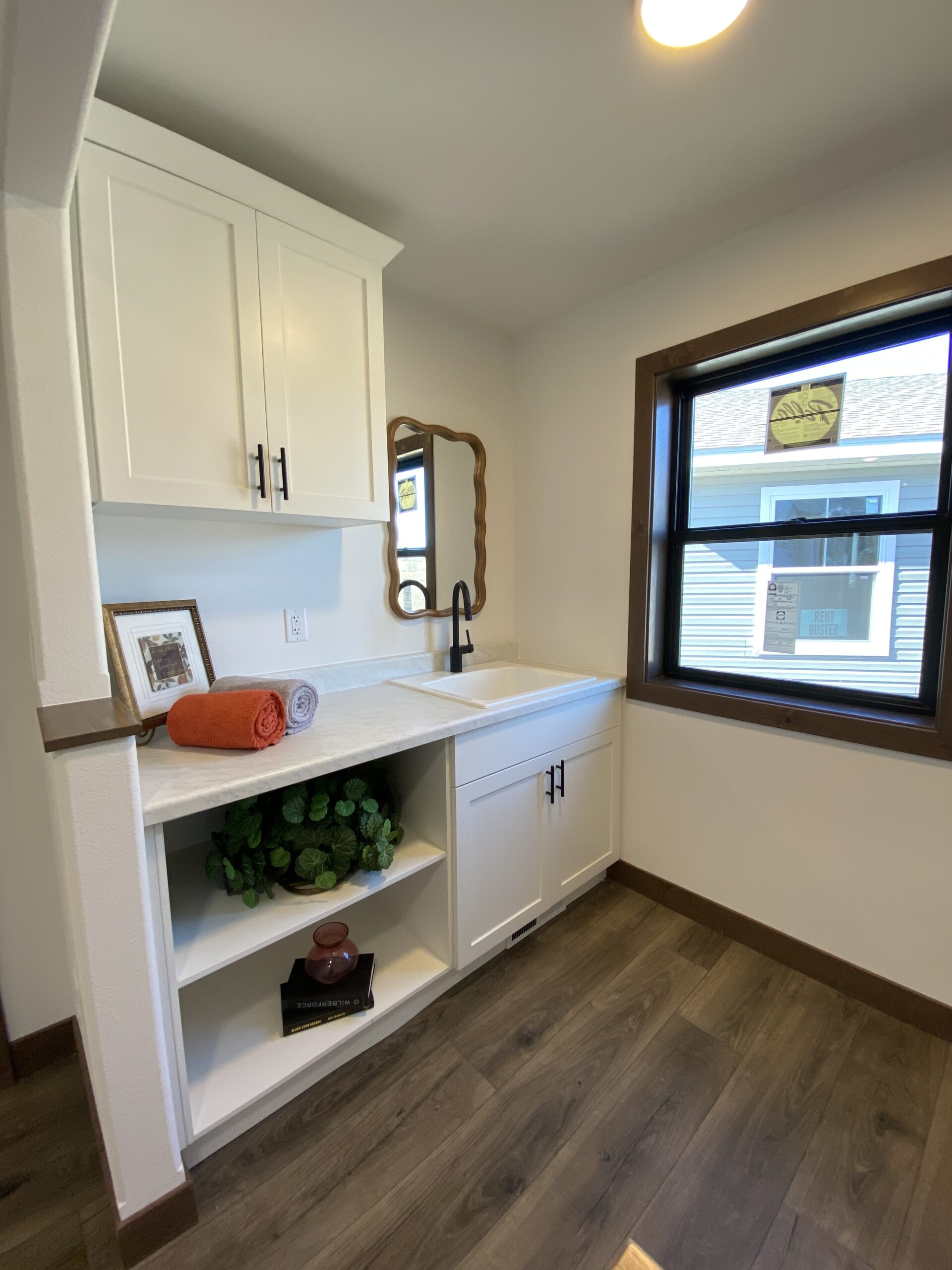 Laundry room with white cabinets, a black faucet on a countertop sink, a window, a shelf with decor items, and a rolled orange towel, creating a tidy and inviting space.