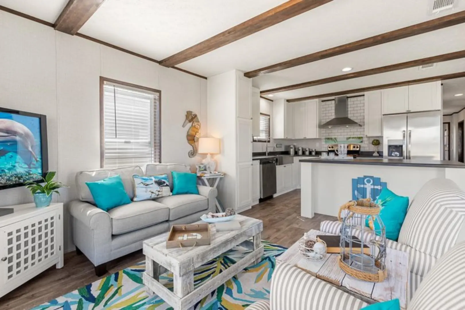 Bright living room with coastal decor, featuring a white couch with turquoise pillows, a TV displaying a dolphin, and a kitchen in the background.