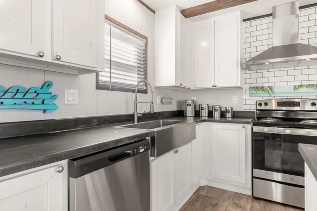 Modern kitchen with white cabinets, stainless steel appliances, and dark countertops. A beach-themed sign and subway tile backsplash add a coastal touch.
