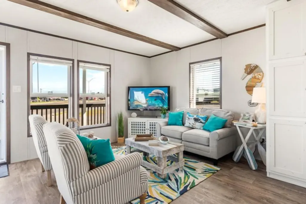 Cozy living room with striped armchairs, a gray sofa, and turquoise cushions. A TV shows ocean scenes. Bright and airy with wooden beams and large windows.