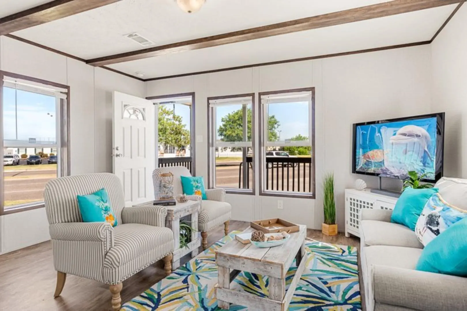 Bright living room with large windows, striped armchairs, and a white sofa adorned with blue ocean-themed pillows. A TV displays aquatic scenes, and a colorful rug adds vibrancy.