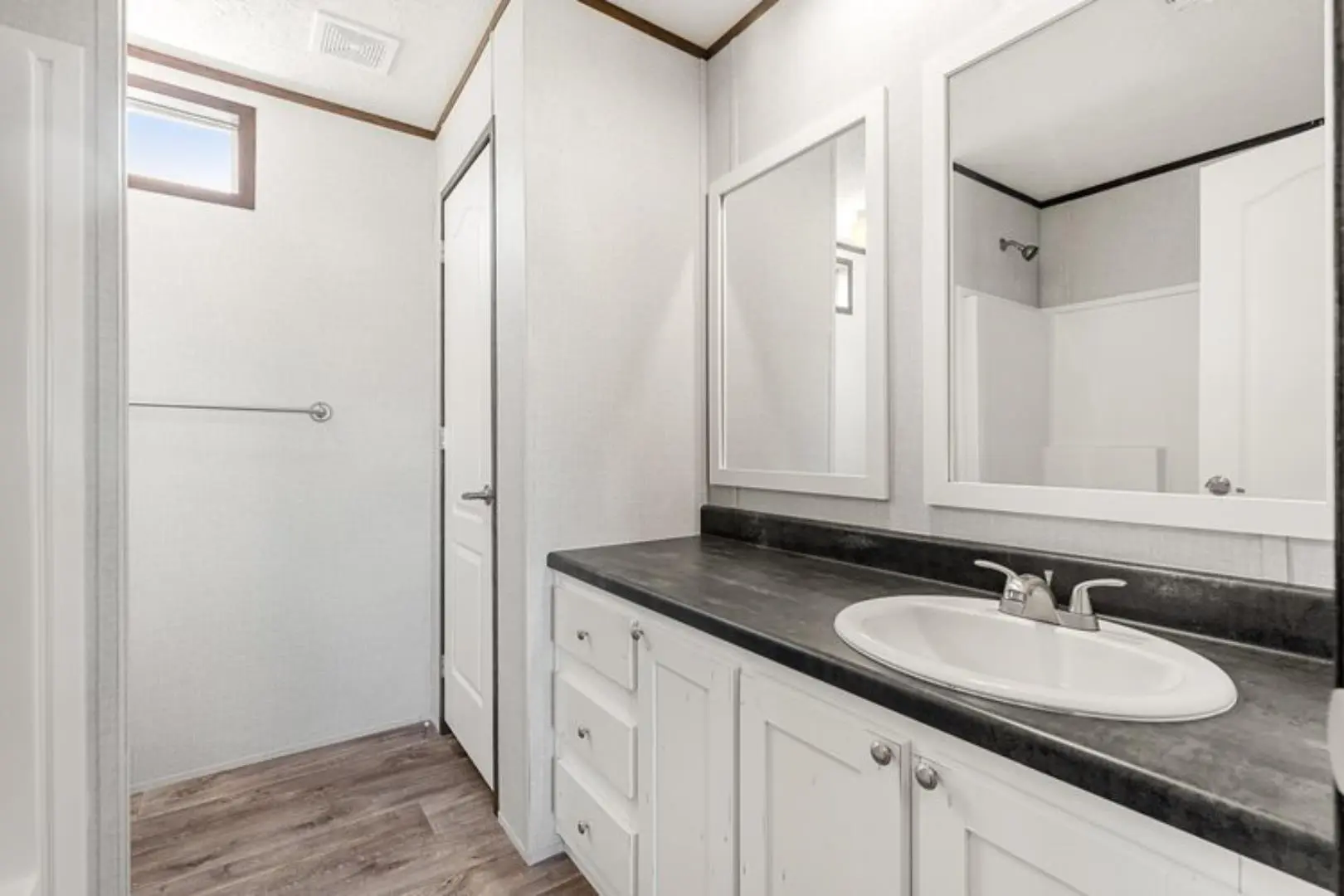 Compact bathroom with light wooden floors, white walls, and a small window. Features include a white vanity with a dark countertop, a sink, and a mirror, creating a clean, minimalist feel.