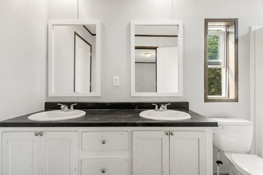 Dual-sink bathroom with black countertop and white cabinets. Two mirrors above sinks. A small window on the right lets in natural light. Calm, neutral tones.
