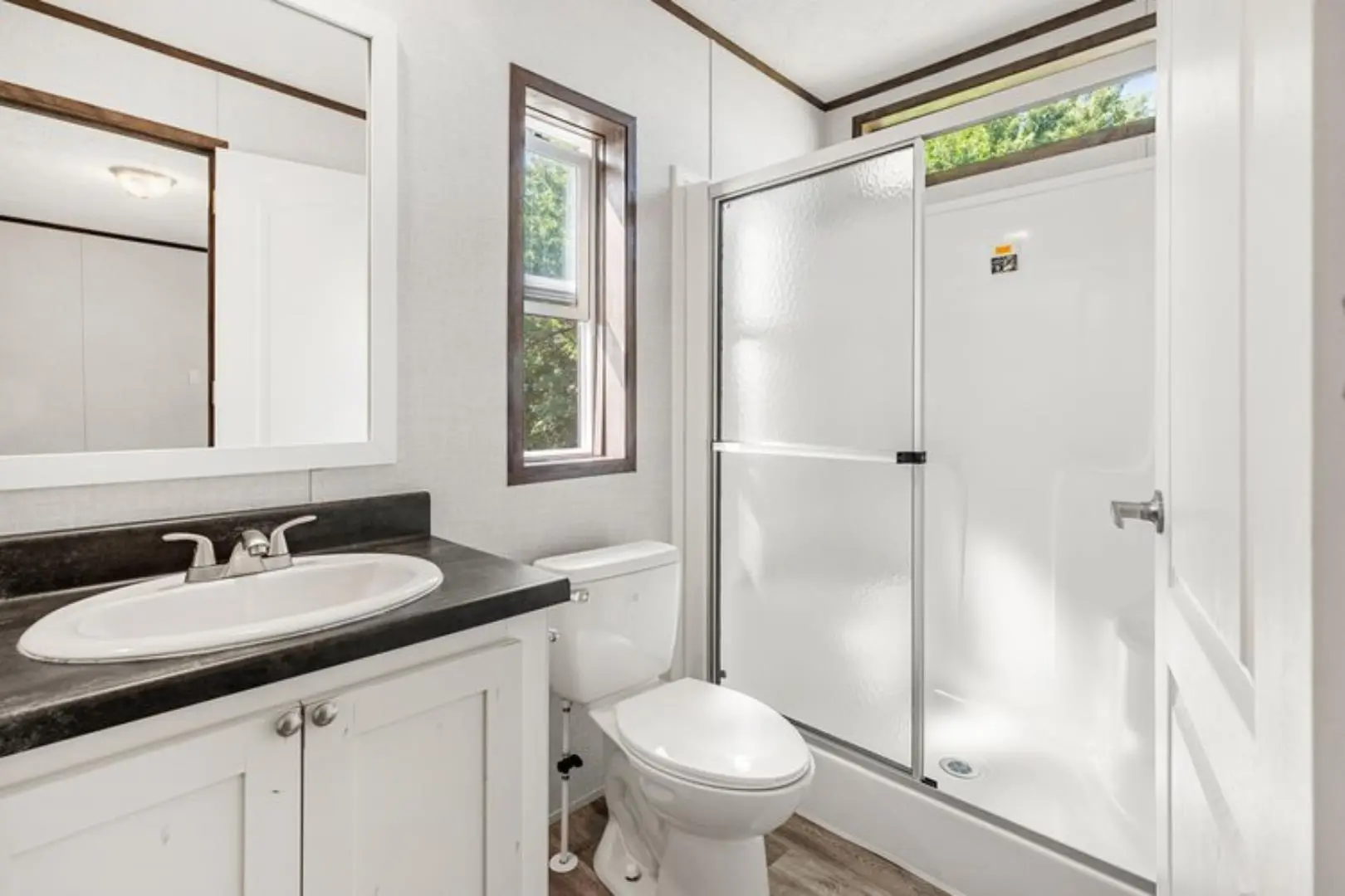 Bright, clean bathroom with white sink, black countertop, and simple cabinetry. A frosted glass shower door, natural light from windows adds warmth.