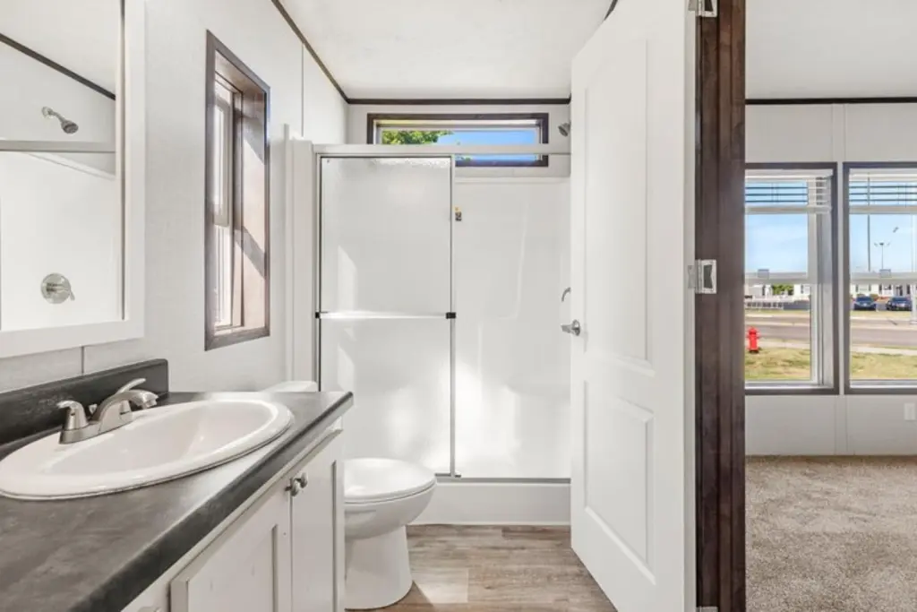 Bright, modern bathroom with a large window. Features a white vanity, black countertop, and a shower next to a toilet. Adjacent room has carpet flooring.