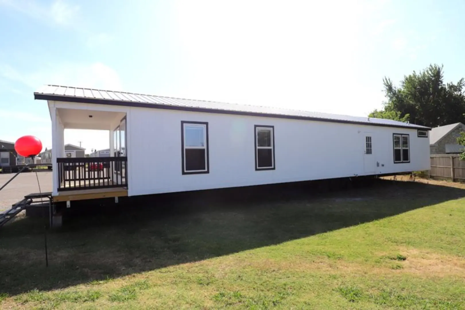 A sleek white mobile home set against a bright sky. It rests on grassy land, with a covered porch and red balloon, suggesting a welcoming atmosphere.