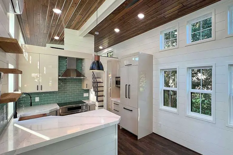 Modern kitchen with white cabinets, green tile backsplash, and dark wood ceiling. Sunlight streams through windows, creating a warm, cozy ambiance.