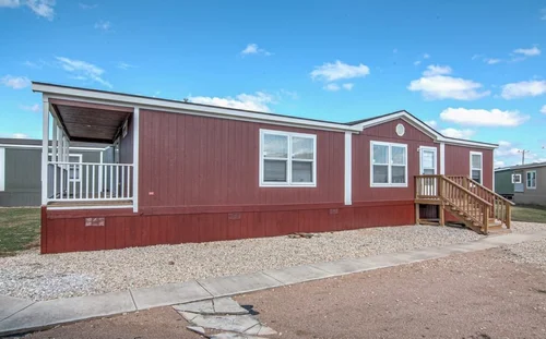 A red mobile home with white trim, a covered porch, and wooden steps is set against a clear blue sky, creating a bright and welcoming scene.