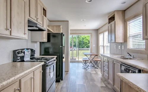 Bright kitchen with wood cabinets, black appliances, and light countertops. A small dining table by a window with greenery outside. Airy and inviting.