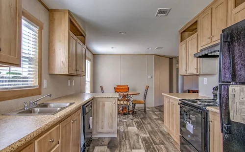A modern kitchen with wooden cabinets, stainless steel sink, and black appliances. Light wood flooring leads to a bright dining area with natural light.