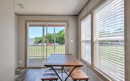 Bright room with a wooden table and benches, large windows with blinds, and sliding glass doors leading to a porch overlooking a grassy area and trees.