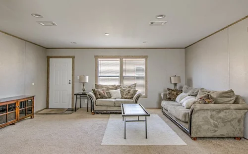 A cozy living room with light beige walls, two plush sofas around a glass coffee table, a wooden TV stand on the left, and soft overhead lighting.