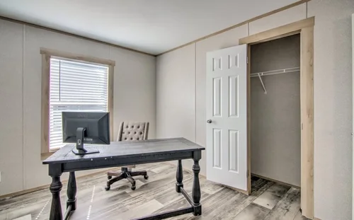 A minimalist home office with a dark wooden desk, computer monitor, and padded chair on a wood floor. An open closet and window with blinds complete the scene.
