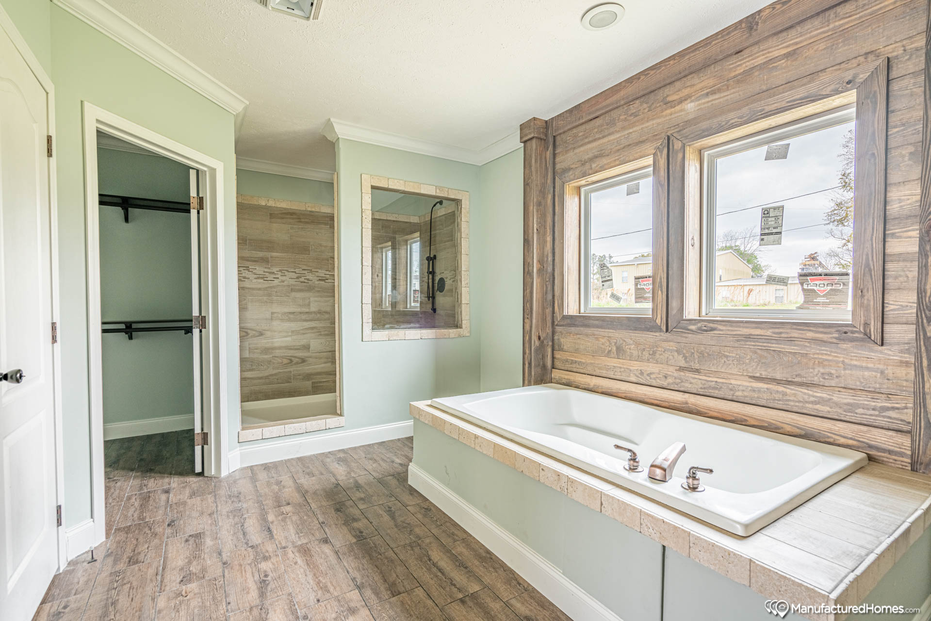 Modern bathroom with wood accents, featuring a large tub beneath two windows, a walk-in shower, and a closet. Natural light and a calming ambiance.