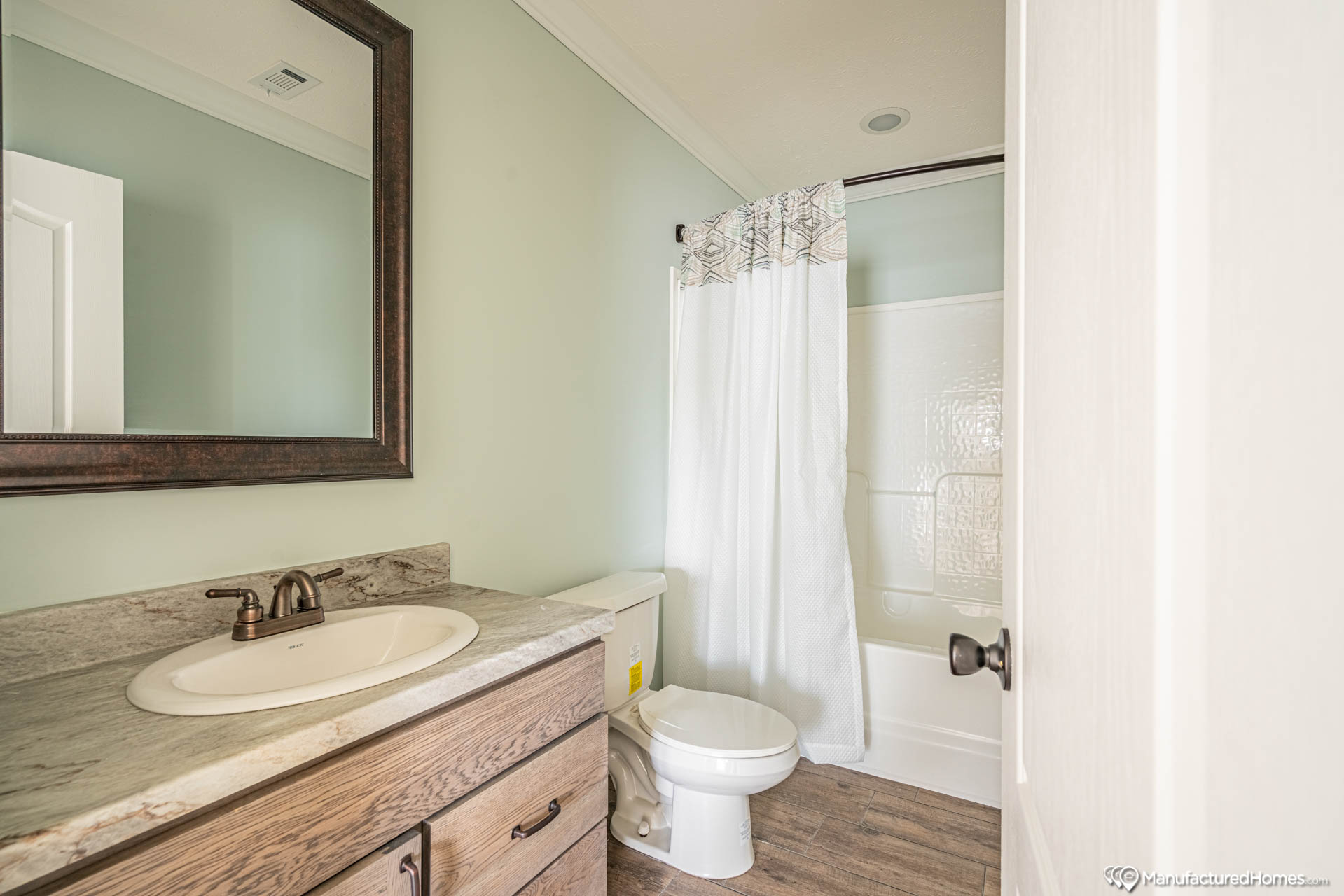 A minimalist bathroom with light green walls features a wooden vanity with a marble countertop, a dark-framed mirror, and a white shower curtain.