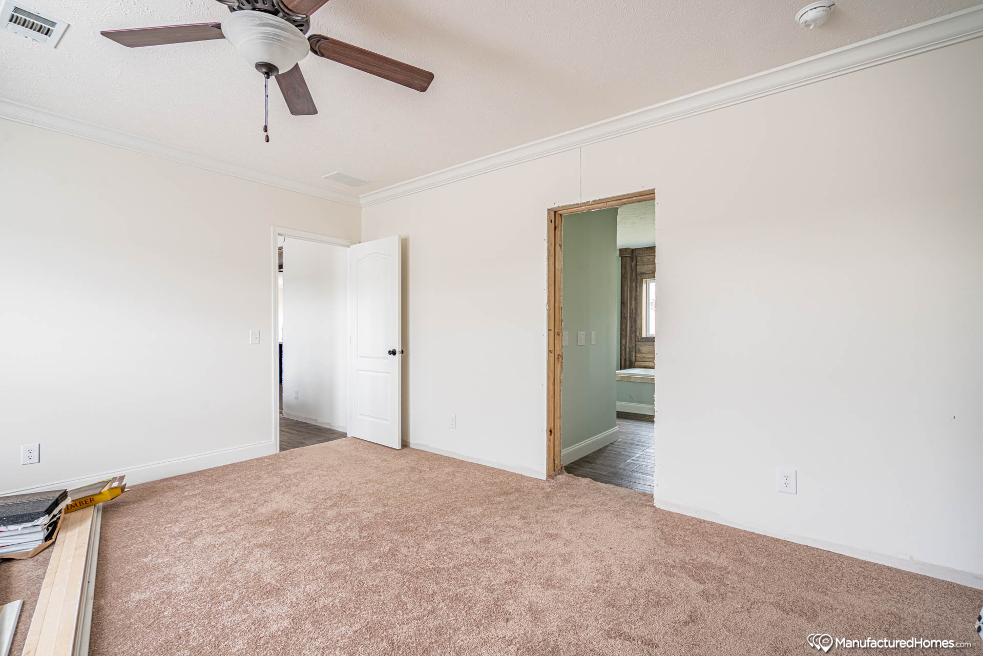 A partially furnished room with beige carpet, a white ceiling fan, and two doorways. One doorway leads to another room with visible wood flooring.