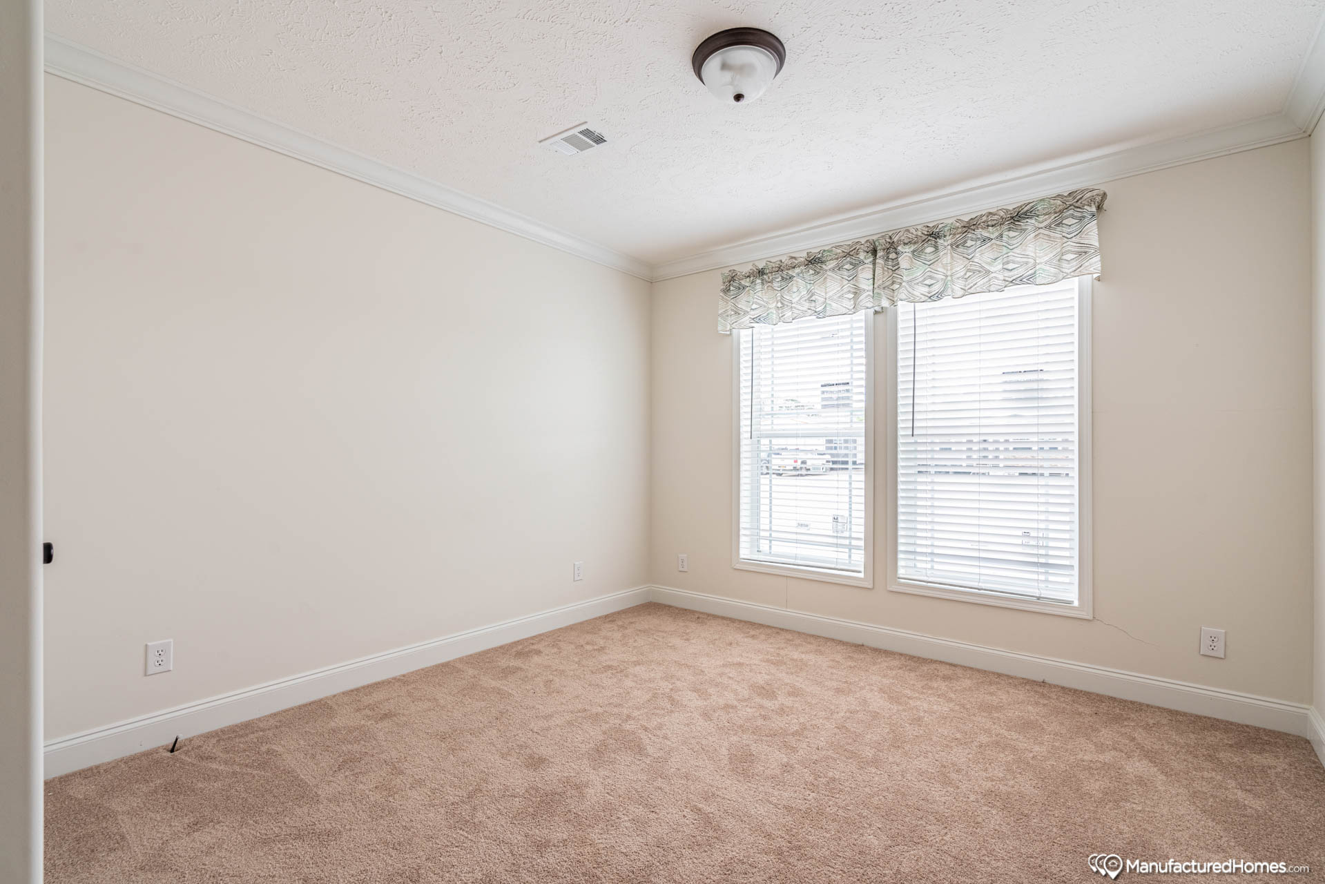 An empty room with beige carpet, cream walls, and a ceiling light. Two large windows with sheer, patterned valances allow natural light to enter.
