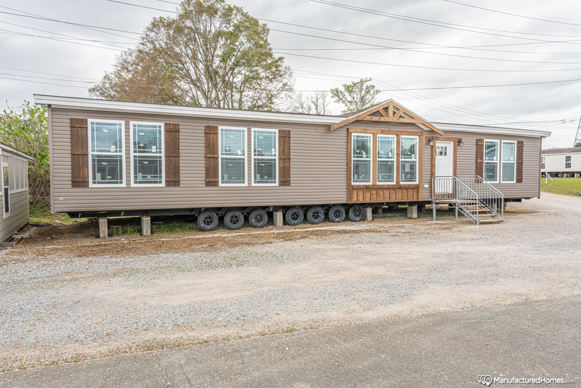 A manufactured home on wheels with brown siding and white trim, featuring large windows and wooden shutters, parked on a gravel lot, with an overcast sky.