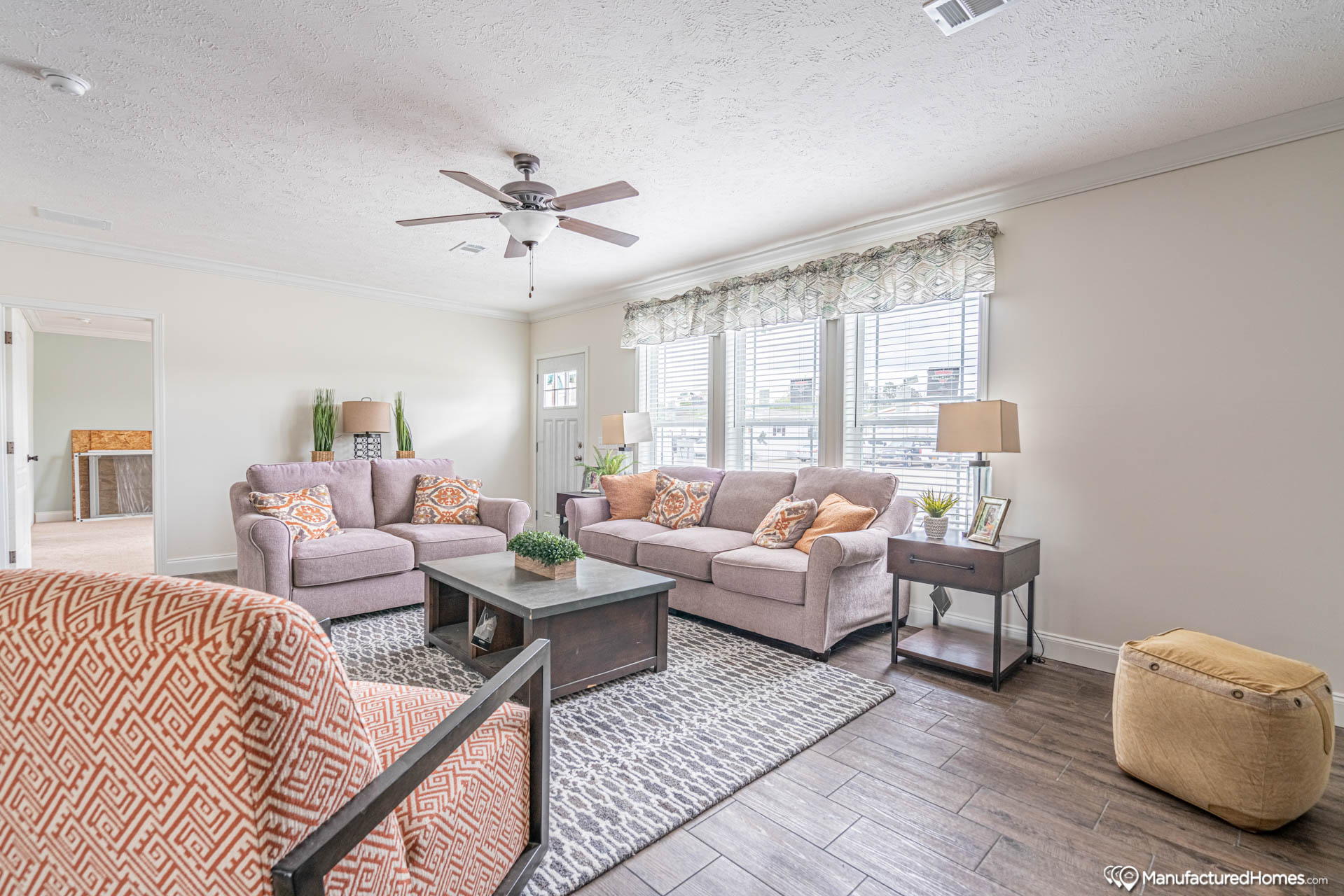 Bright living room with pastel sofas and decorative pillows, a geometric armchair, and a central coffee table. A ceiling fan and large windows add light.