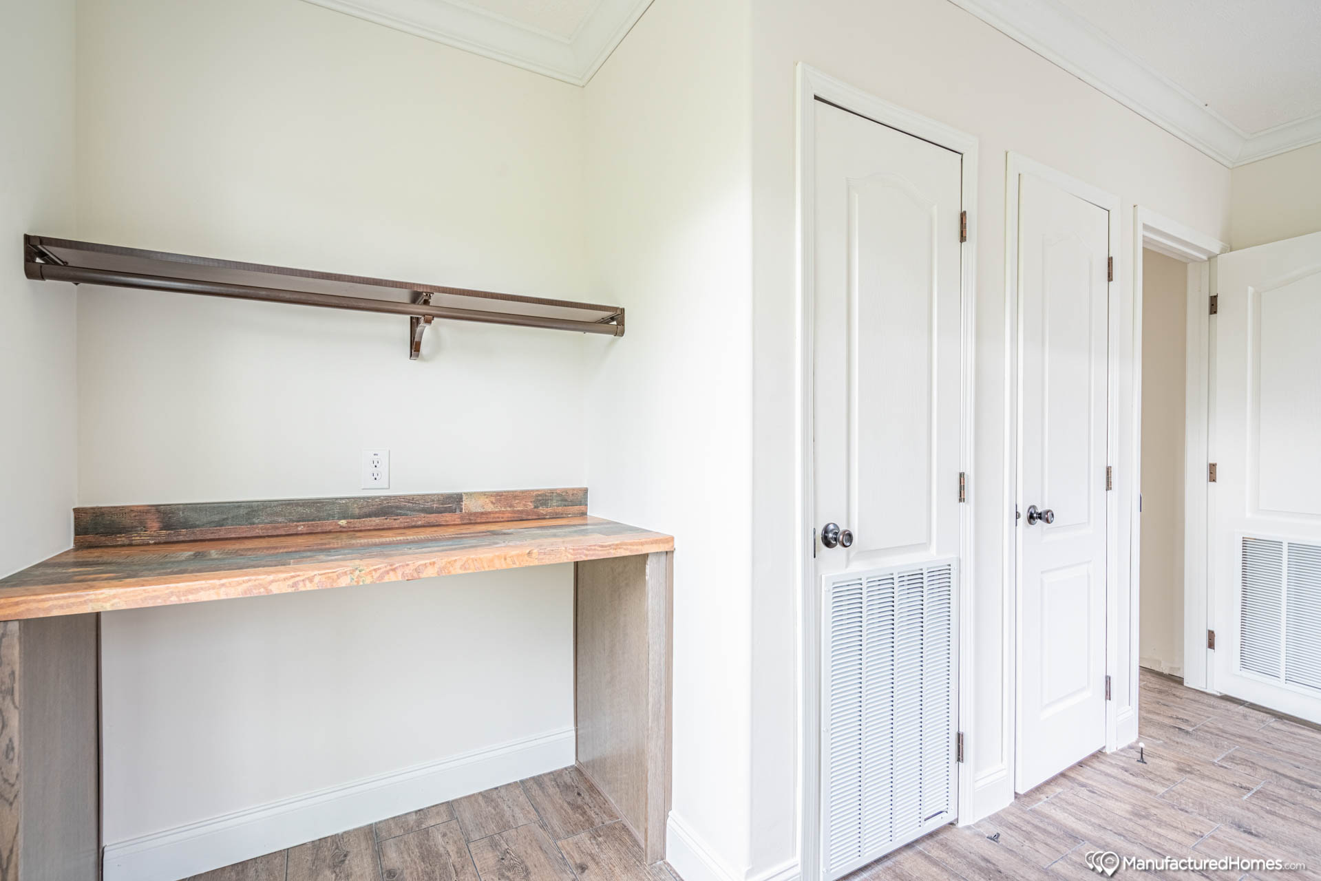 A compact, minimalist workspace features a wooden desk and a metal shelf against a cream wall. Two white doors are nearby. The setting feels clean and modern.