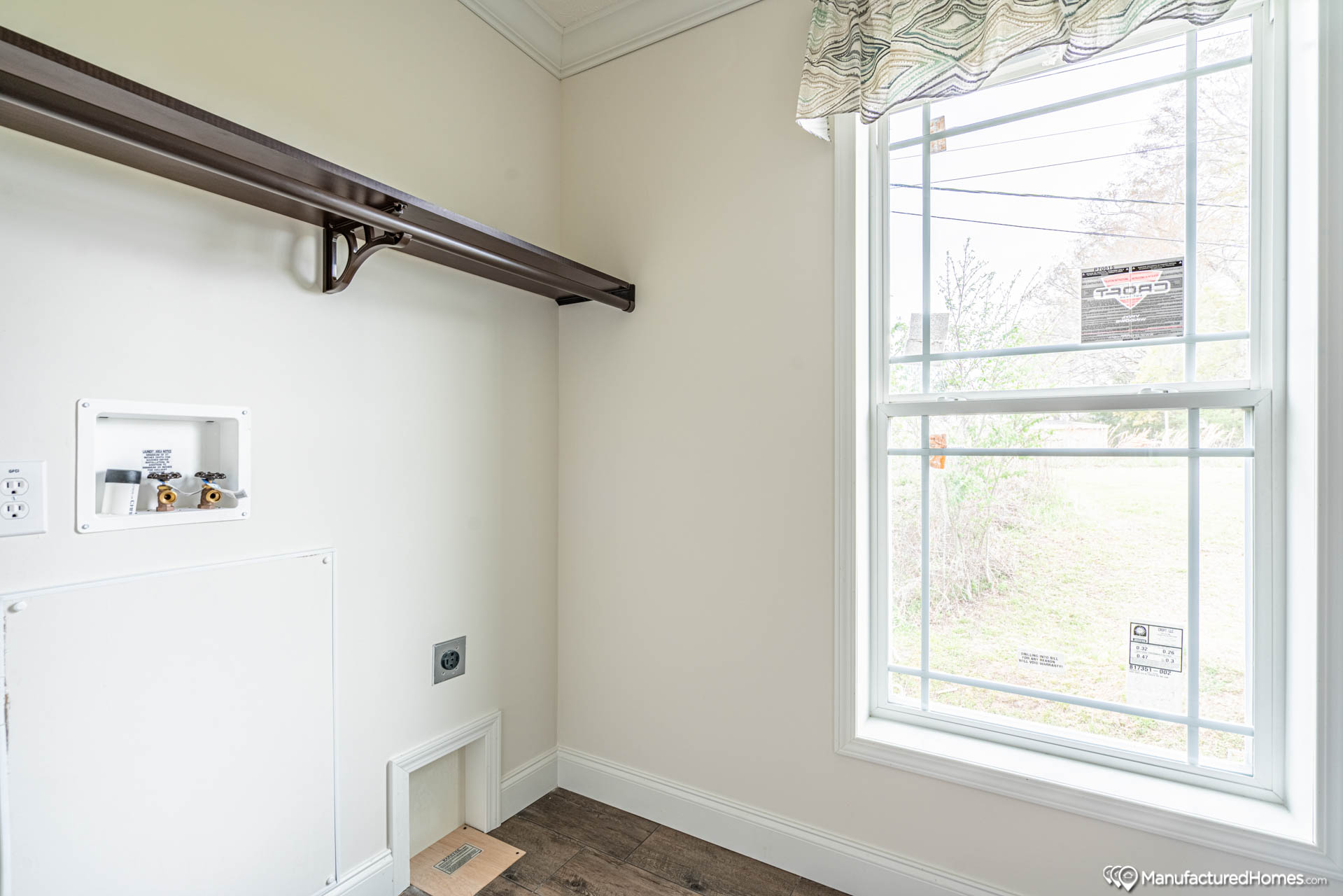 A small laundry room with a wooden shelf and plumbing connections for a washer under bright window light. Hardwood floor and minimal decor.