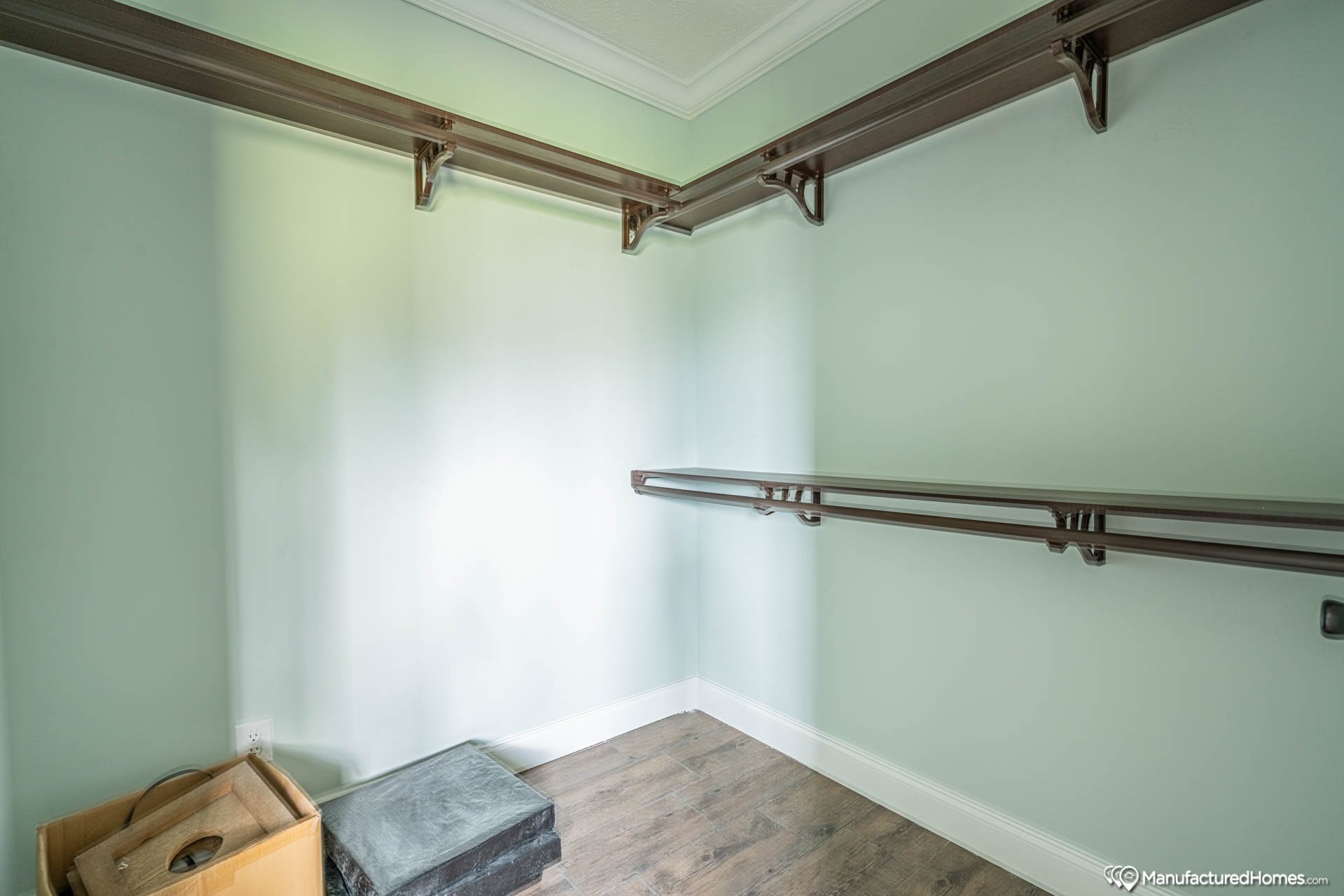 Spacious, empty walk-in closet with light green walls and wooden floor. Shelving and rods line the upper walls. A cardboard box and a cushion are on the floor.
