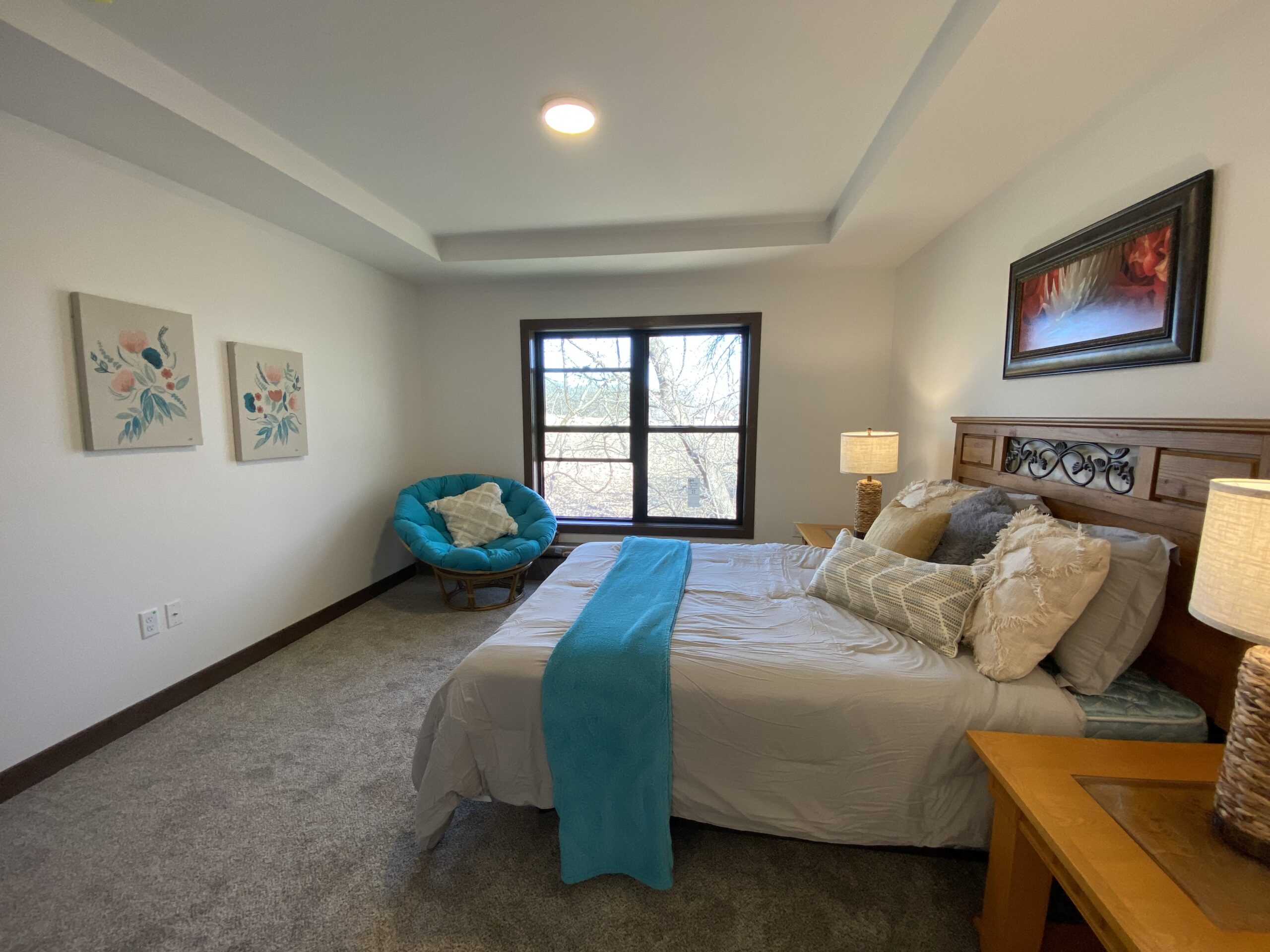 A cozy bedroom with a queen bed adorned with white and turquoise bedding. A round turquoise chair and floral artwork decorate the serene, well-lit space.