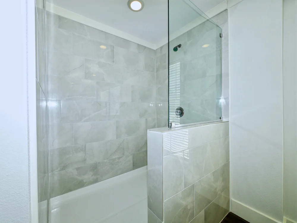 A modern bathroom shower with muted gray tiles and a glass door. The room is bright, creating a clean and minimalist ambiance.