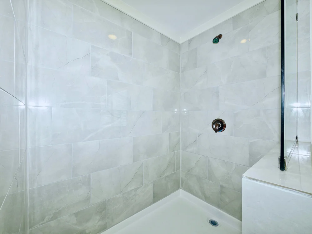 Spacious shower with light gray marble tiles, featuring a dark showerhead and faucet. Glass partition on the right adds modern elegance.