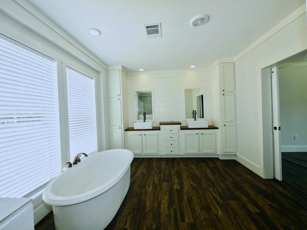 Spacious bathroom with a freestanding oval bathtub, wooden floors, dual sinks and mirrors, white cabinetry, and large windows with blinds.