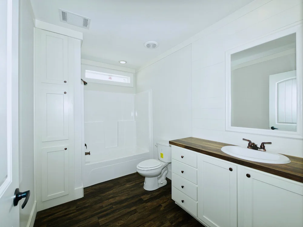 Bright, modern bathroom with white cabinets and walls, wooden countertop, and dark wood floor. Contains a sink, bathtub-shower combo, and toilet. Cozy and clean.