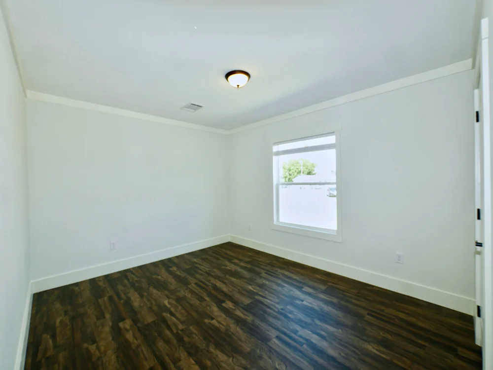 A bright, empty room with white walls and dark wooden floors. A single window on the right lets in natural light, while a ceiling light adds warmth.