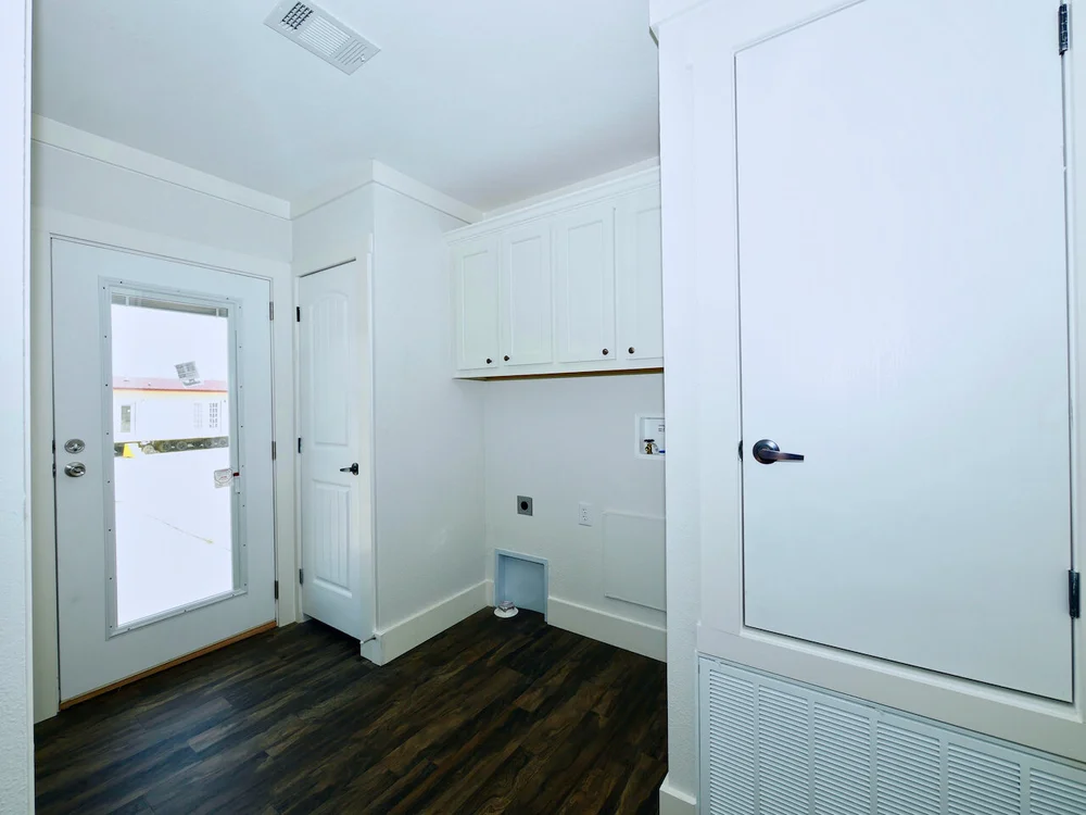 Bright laundry room with white walls and cabinets, dark wood flooring, and a door with a large glass panel. The space feels clean and minimalistic.