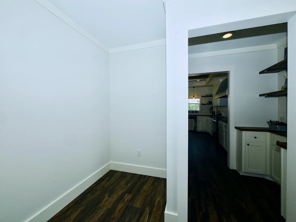 Empty white-walled room with dark wooden floor, leading to a kitchen through an open doorway. The kitchen has dark flooring, white cabinets, and shelves.