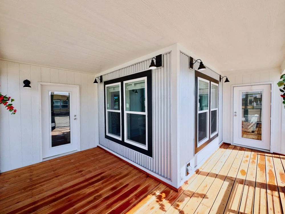 Bright wooden deck corner, bordered by white paneled walls and black-framed windows. Two doors lead inside, with potted plants adding a touch of color.