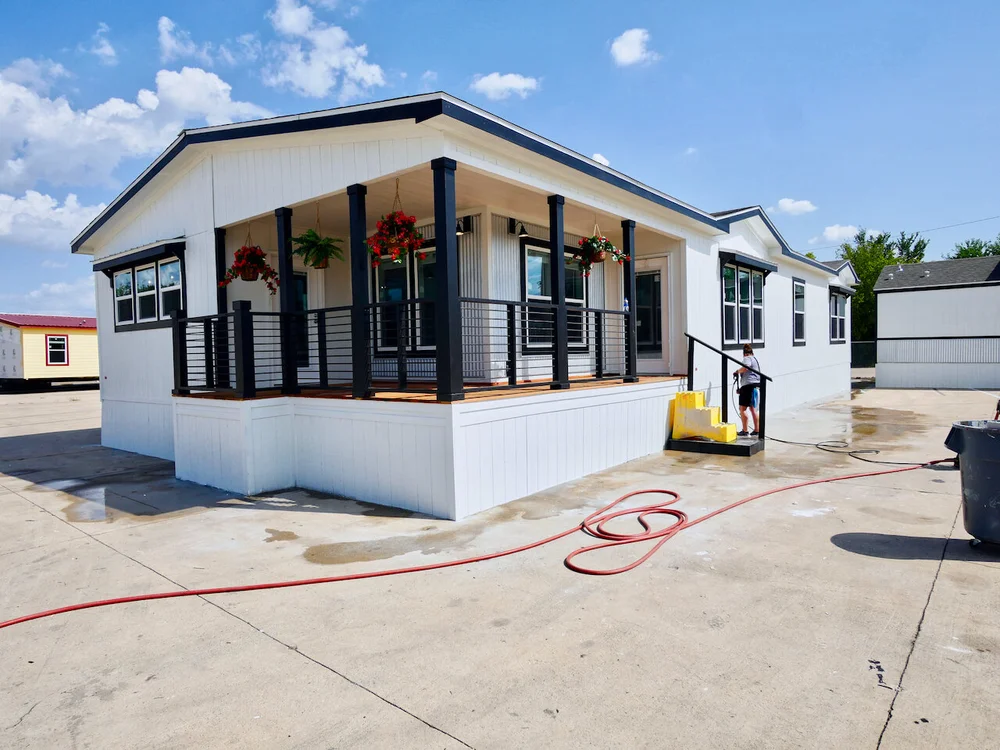 Modern modular home with a covered porch, black railings, and hanging plants. A child stands on yellow steps. Bright day, clear sky.