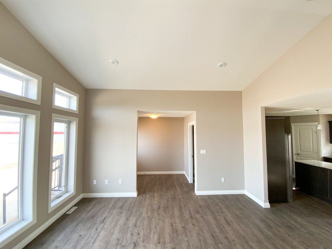 Spacious, empty room with beige walls and wood floors. Large windows on the left let in natural light. Open doorway leads to kitchen with visible appliances.