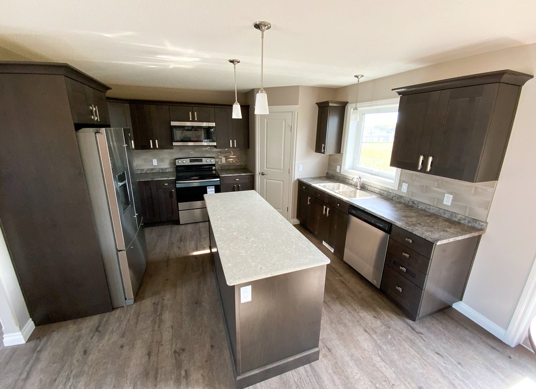 Modern kitchen with dark wood cabinets, a beige marble island, and stainless steel appliances. Sunlight streams through a window, creating a warm atmosphere.