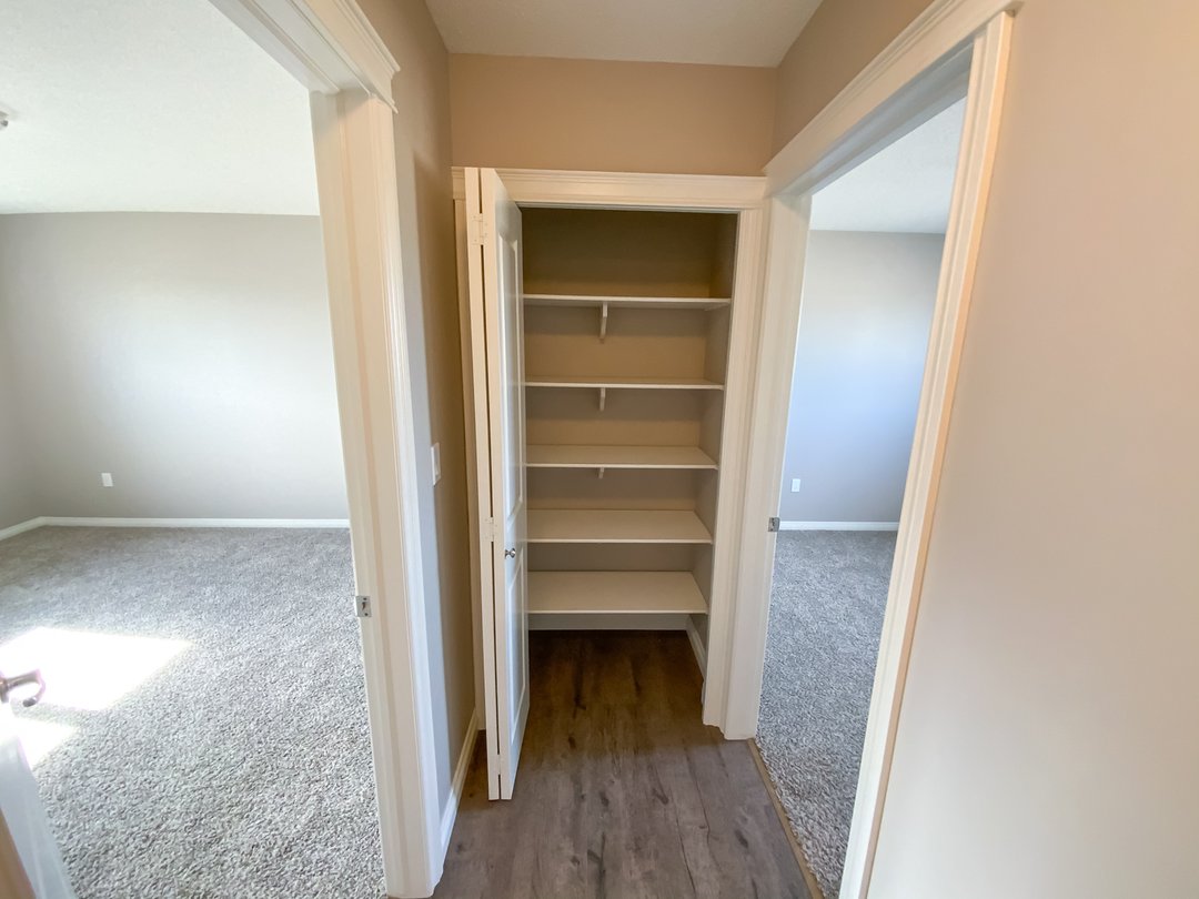 Hallway with two open doors leading to carpeted rooms. A closet with white shelves is centered, and light wood flooring creates a bright, tidy ambiance.