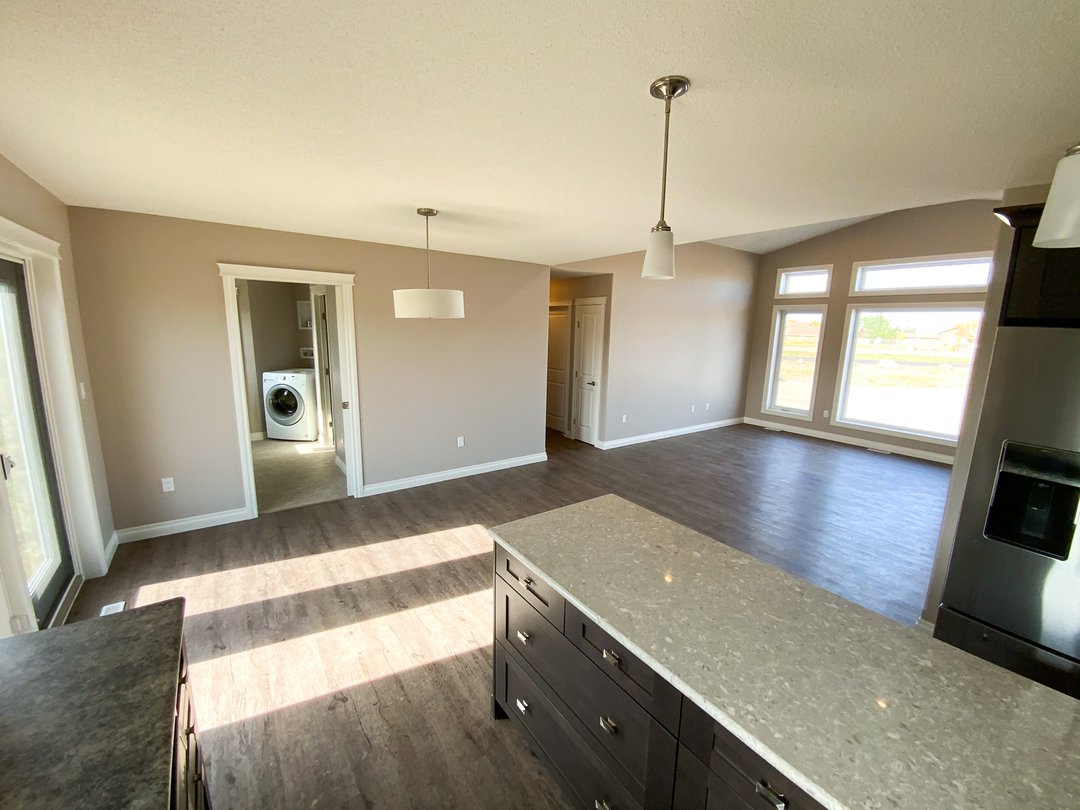 Open floor plan with wood flooring featuring a kitchen island, hanging lights, and large windows. A washer is visible in an adjacent room.