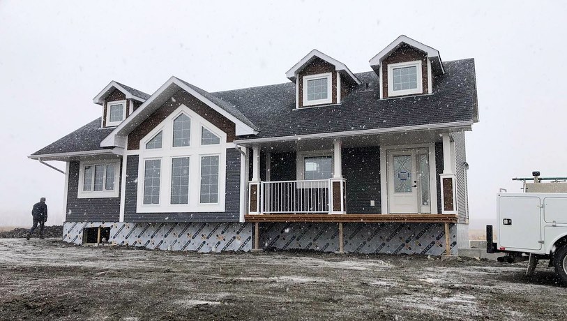 A modern, snow-dusted house with a gray exterior and white trim. It features large windows and dormers. A person and a white truck are nearby.