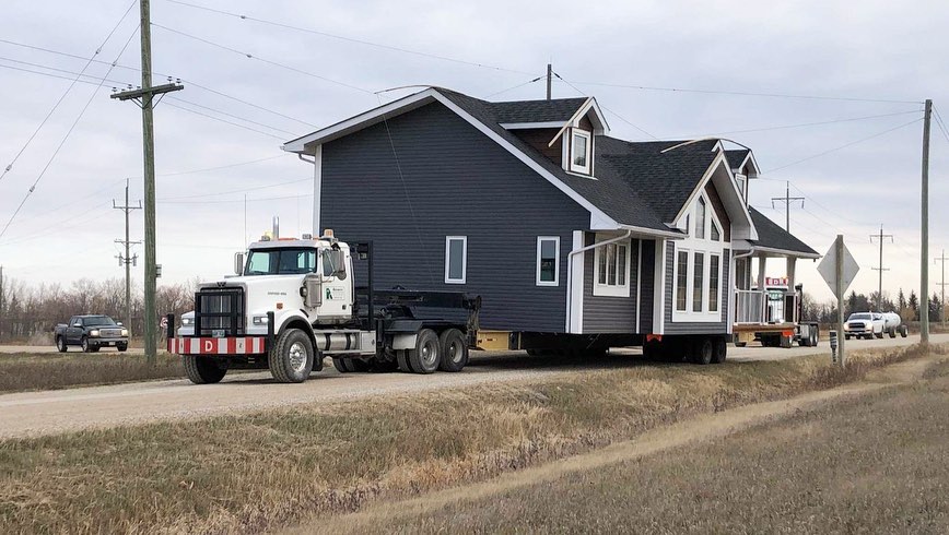 A large house is being transported by a flatbed truck on a rural road. The house is dark gray with white trim, and the sky is overcast.
