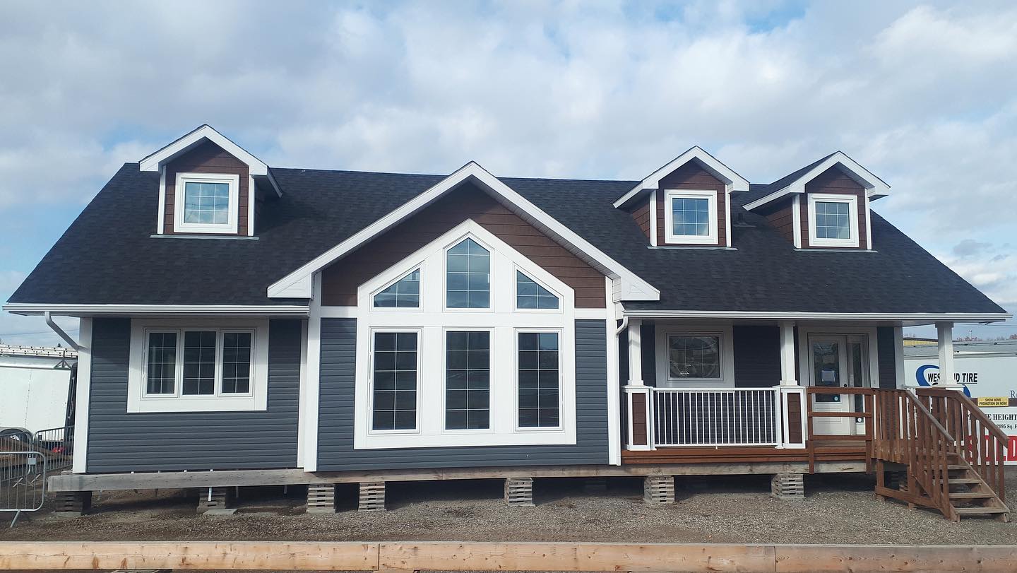 A modern, single-story house with gray siding, white trim, and a dark roof. It has large windows, dormers, and a welcoming front porch set against a cloudy sky.