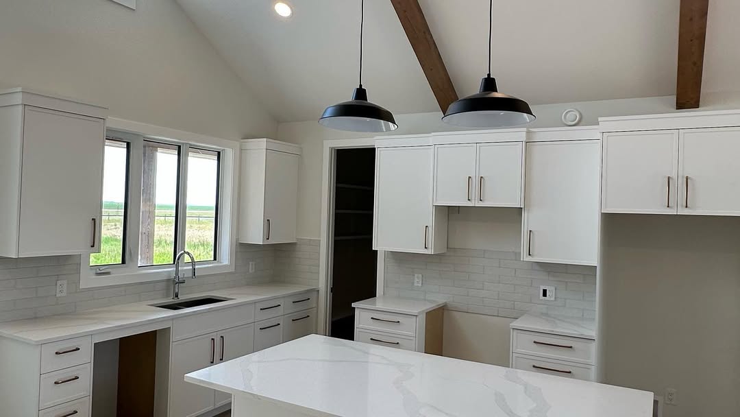 A modern kitchen with white cabinetry, marble countertops, and black pendant lights. Large windows and wooden ceiling beams add warmth and natural light.