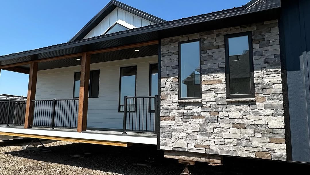 A modern prefab home with a wide porch, featuring wooden pillars, white siding, and stone accents. The house rests on temporary supports, under a clear blue sky.