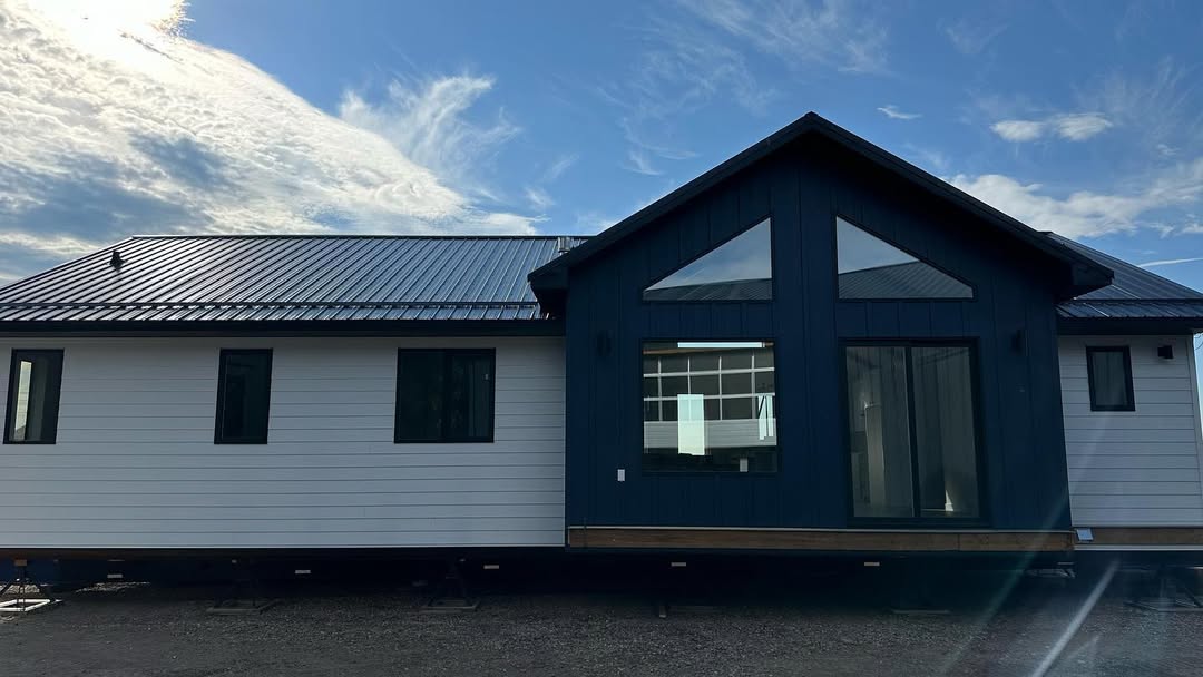A modern house under a bright blue sky with clouds. It features dark and light contrasting siding, large windows, and a pitched metal roof.