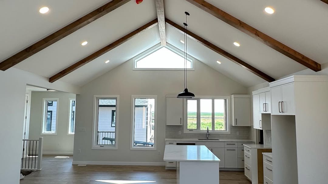 Spacious kitchen with high vaulted ceiling and exposed wooden beams. Bright windows, modern white cabinets, and an island create an airy, welcoming atmosphere.